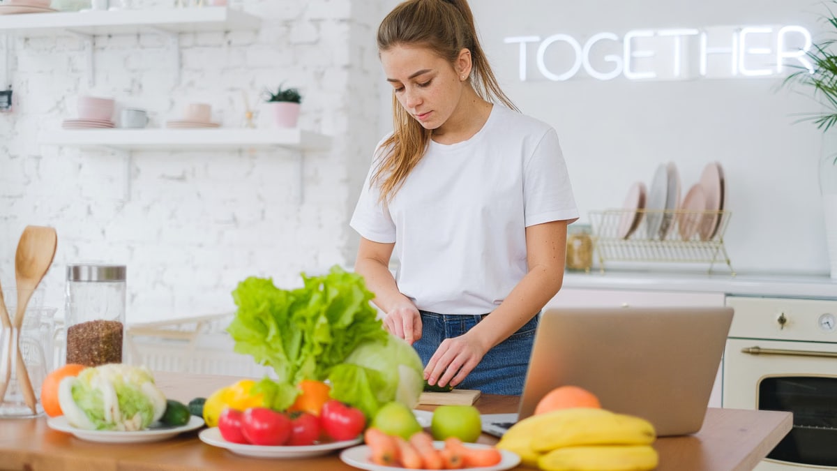 Woman in white t-shirt preparing fresh vegetables in a bright kitchen with a laptop.