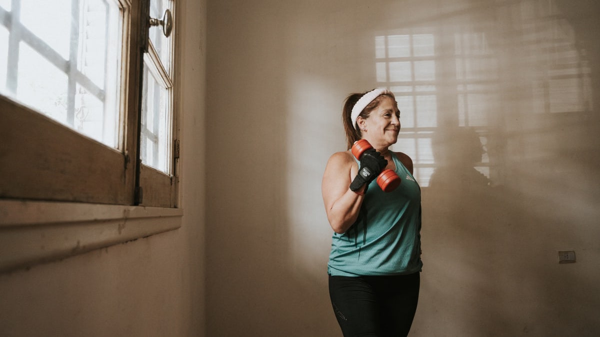 A woman lifting dumbbells indoors by a window, focused on her workout routine.